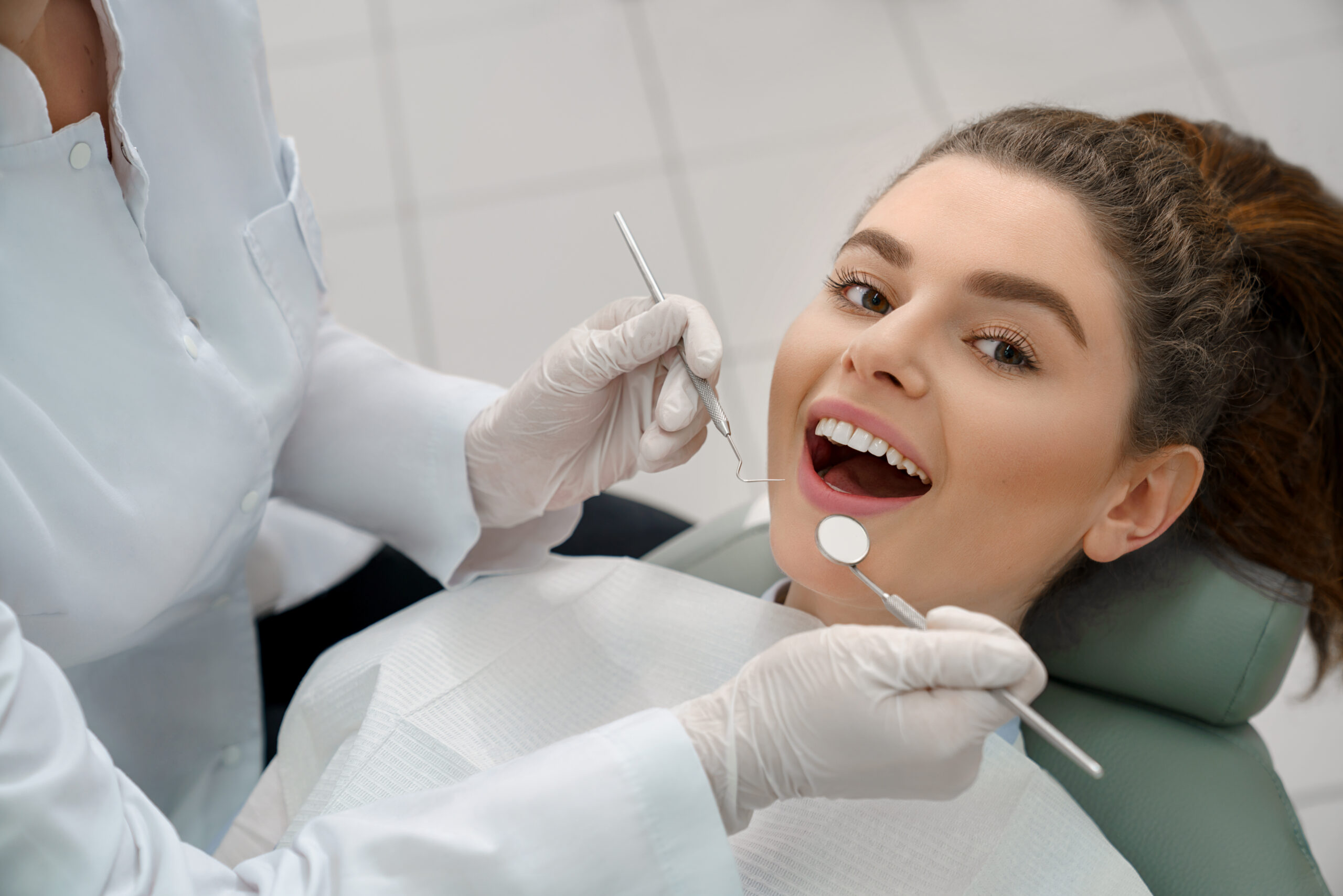 View from above of young woman lying on dental chair with open mouth and looking at camera while professional doctor checking teeth. Concept of dentistry procedure and stomatology.