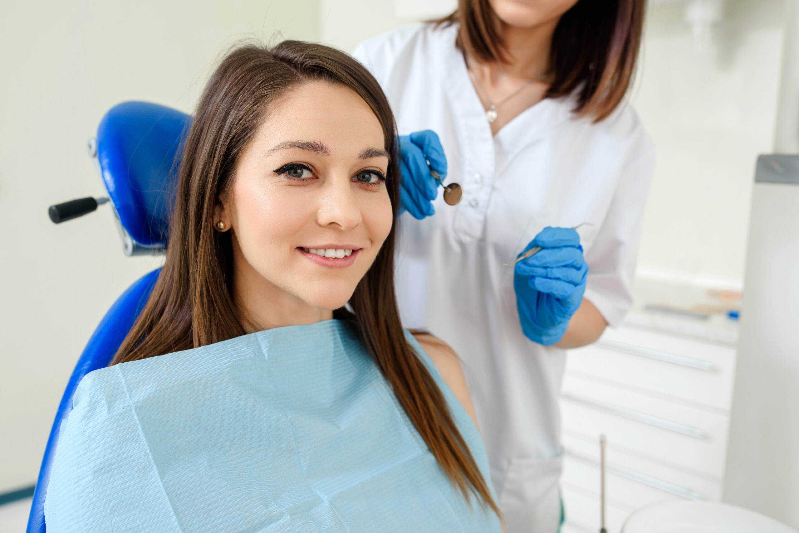 Young happy smiling woman patient in a dental clinic is receiving a dental treatment and looking at the camera. The dentist with dental equipment are behind