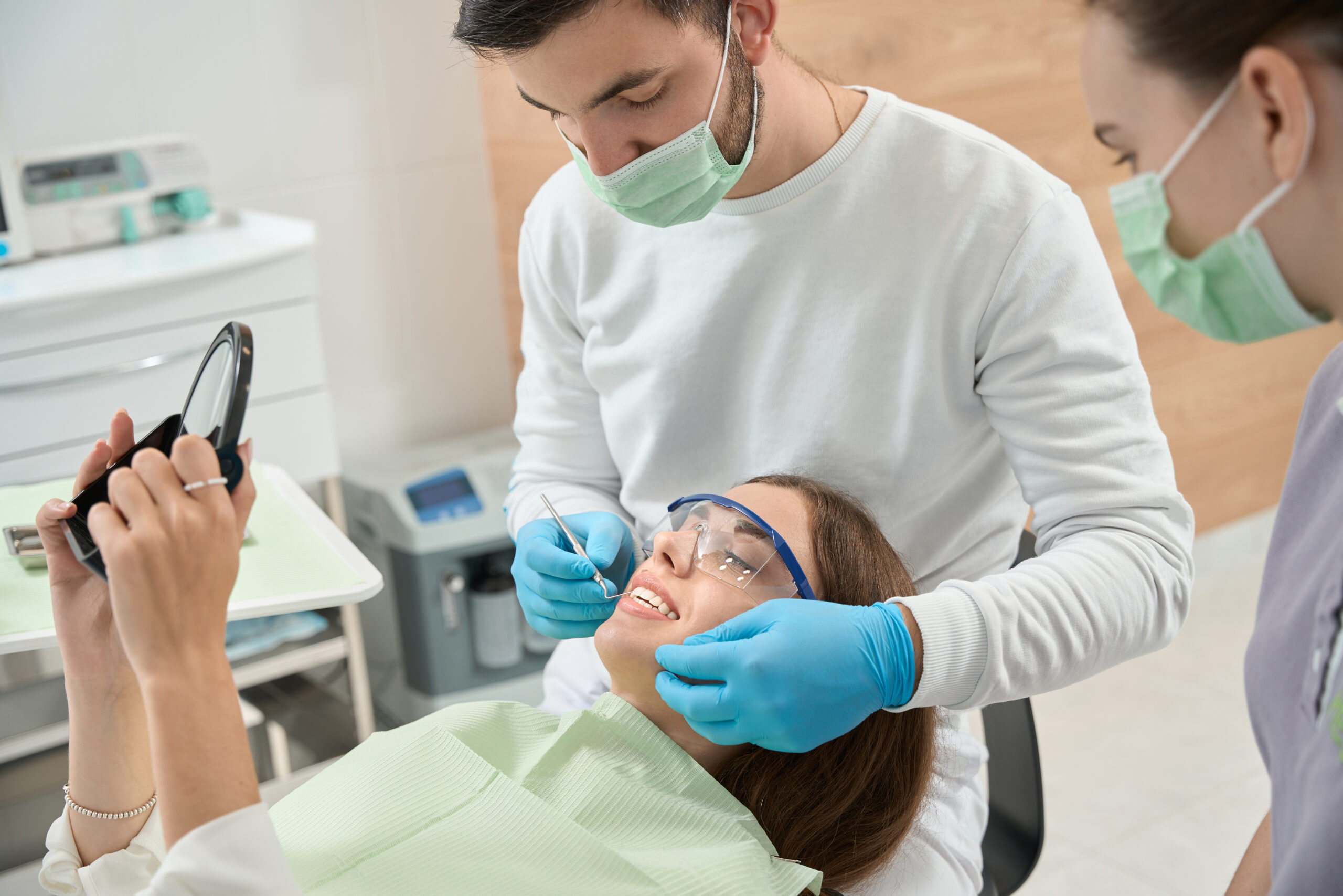 Woman looking in hand mirror while dentist checking her anterior teeth with dental probe in presence of nurse